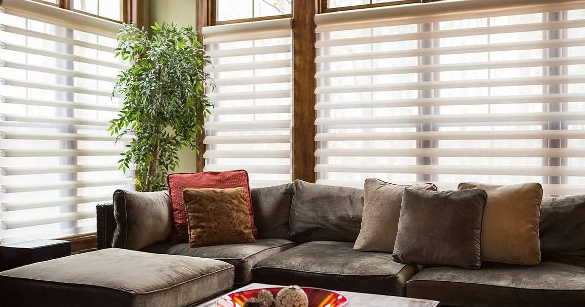 Modern living room with comfortable sectional sofa, soft pillows, and large windows with white blinds.