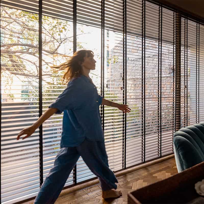 Relaxed woman enjoying morning sunlight through window blinds at home.